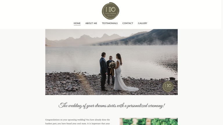A bride and groom stand by a mountain lake, holding hands during an outdoor wedding ceremony with an officiant. The scene is calm, with mist over the water and mountains in the background. Website navigation is visible above the photo.